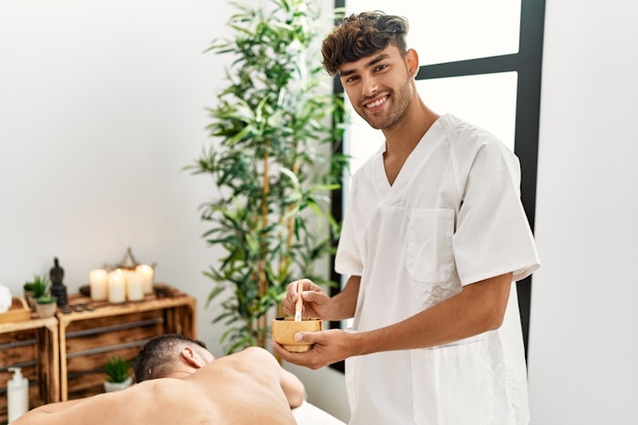 Two hispanic men physiotherapist and patient having skin back treatment using exfoliating lotion at spa