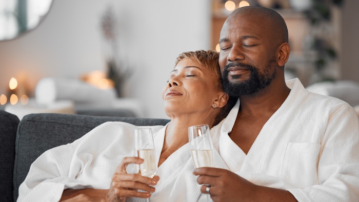 Mature couple, bonding and champagne on spa date, romantic retreat or resort holiday for marriage anniversary celebration. Black woman, man and alcohol glass on relax salon or hotel living room ...