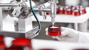 Cropped shot of factory worker in gloves and gown holding plastic jar, filling it with white face or body cream. Beauty, skincare and cosmetology