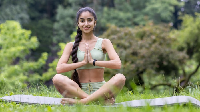 A portrait of a young Indian woman sitting in a park on a rug in a lotus pose and deals with yoga.