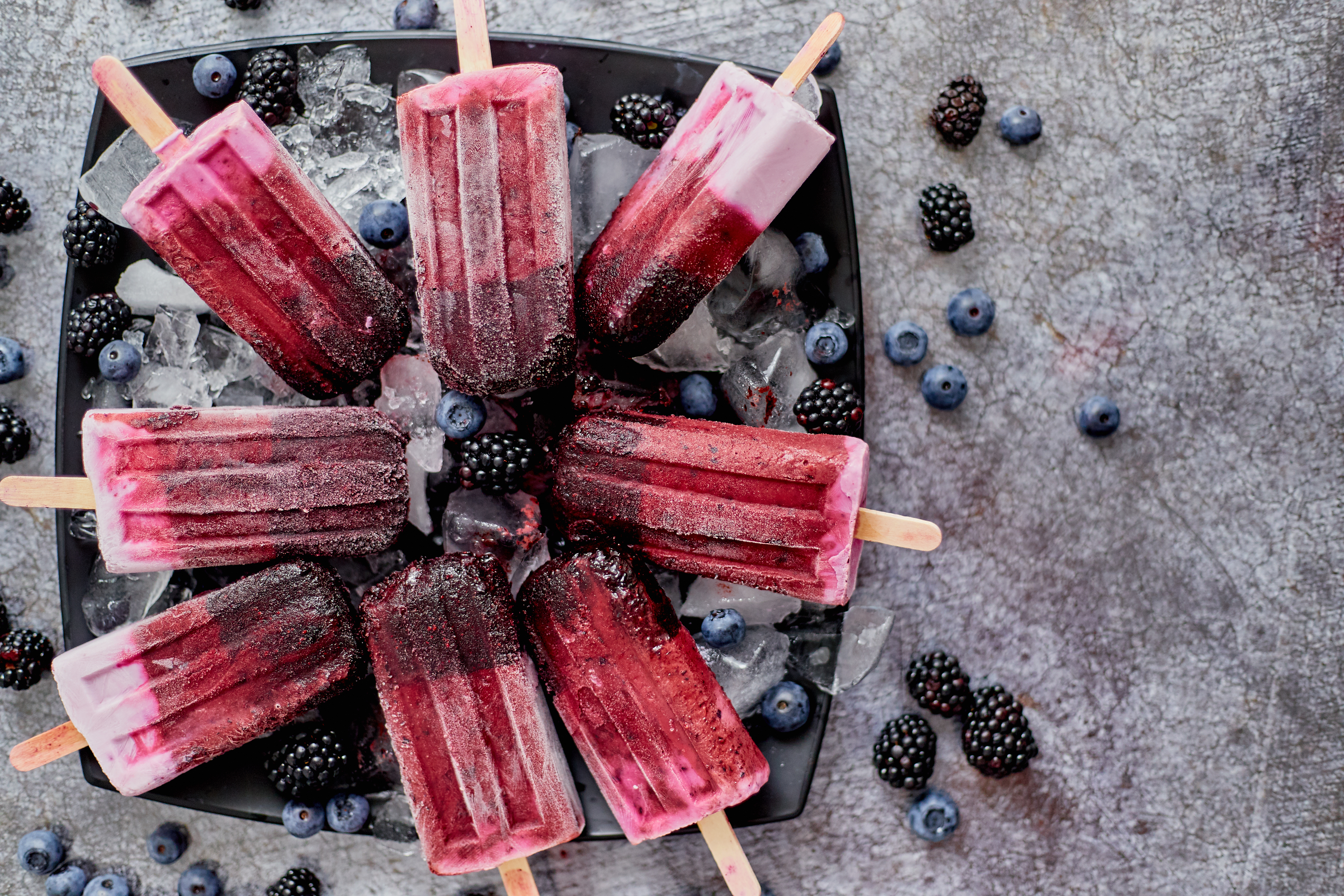 Homemade fresh frozen blueberry and blackberry popsicles on black plate with ice sitting on stone