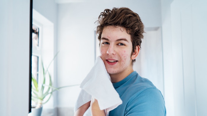 Portrait of smiling teenage boy with acne problem who takes care his face skin at home. He looking in the mirror and wipes face with towel in bathroom.