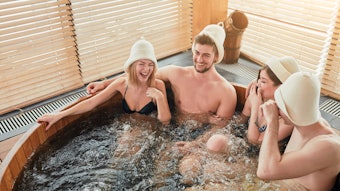 Group of male and female friends visiting bathhouse in holidays, being overjoyed and happy, enjoying jacuzzi in hot wooden round barrel