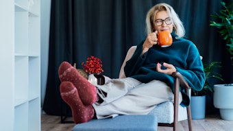 Middle aged woman relaxing with pumpkin shaped cup of hot drink in scandy style hygge interior home with fall mood decor. Lady dreaming, enjoy calm mood without stress, well being alone, self-care concept