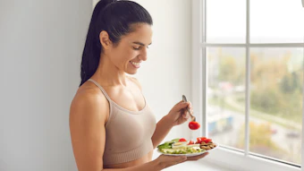 Smiling pretty young fitness woman standing and eating balanced healthy meal before or after workout at home window. Healthy diet, active fitness lifestyle, clean eating food concept