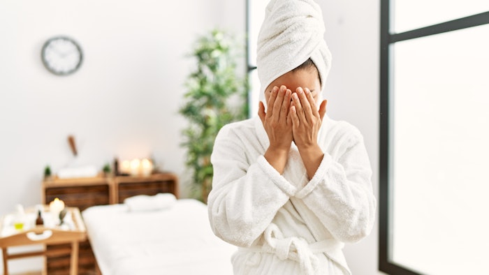 Young brunette woman wearing towel and bathrobe standing at beauty center with sad expression covering face with hands while crying. upset client.