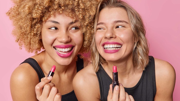 two women smiling wearing pink lipstick holding lipstick tubes