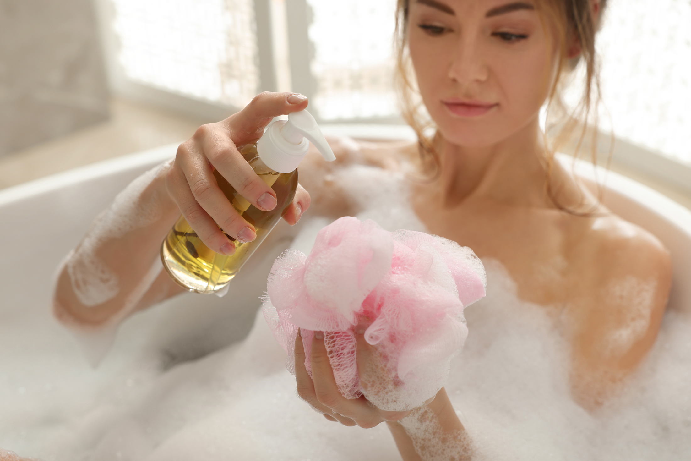 woman in foamy bath pouring body gel into loofah