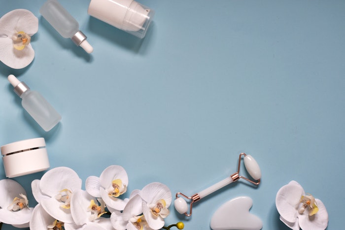 Beauty Products. Facial Roller, essential oils, cream of skincare and beautiful orchids on blue background. Flat lay, top view with copy space. Massage tool for facial skin care, SPA beauty treatment