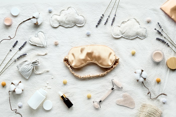 Healthy night sleep flat lay. Pale yellow sleep mask, face stone roller, Gua Sha stone and essential oil. Sleeping pills, night cream. Off white textile bed sheet. Dry lavender, cotton flowers