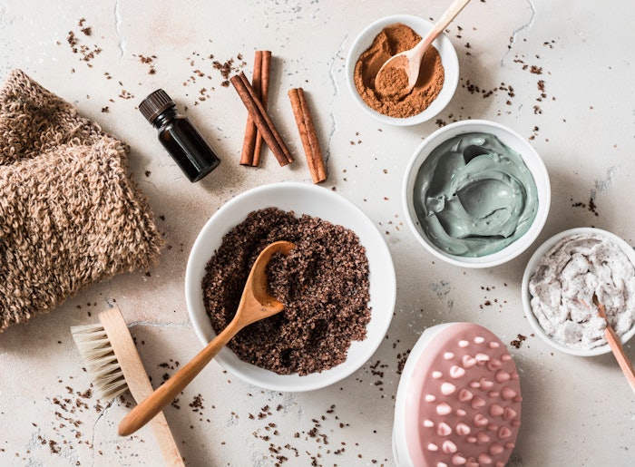 Home anti-cellulite products - coffee scrub, cosmetic clay, essential orange oil, hand anti-cellulite massager, nut scrub, cinnamon, natural loofah on light background.