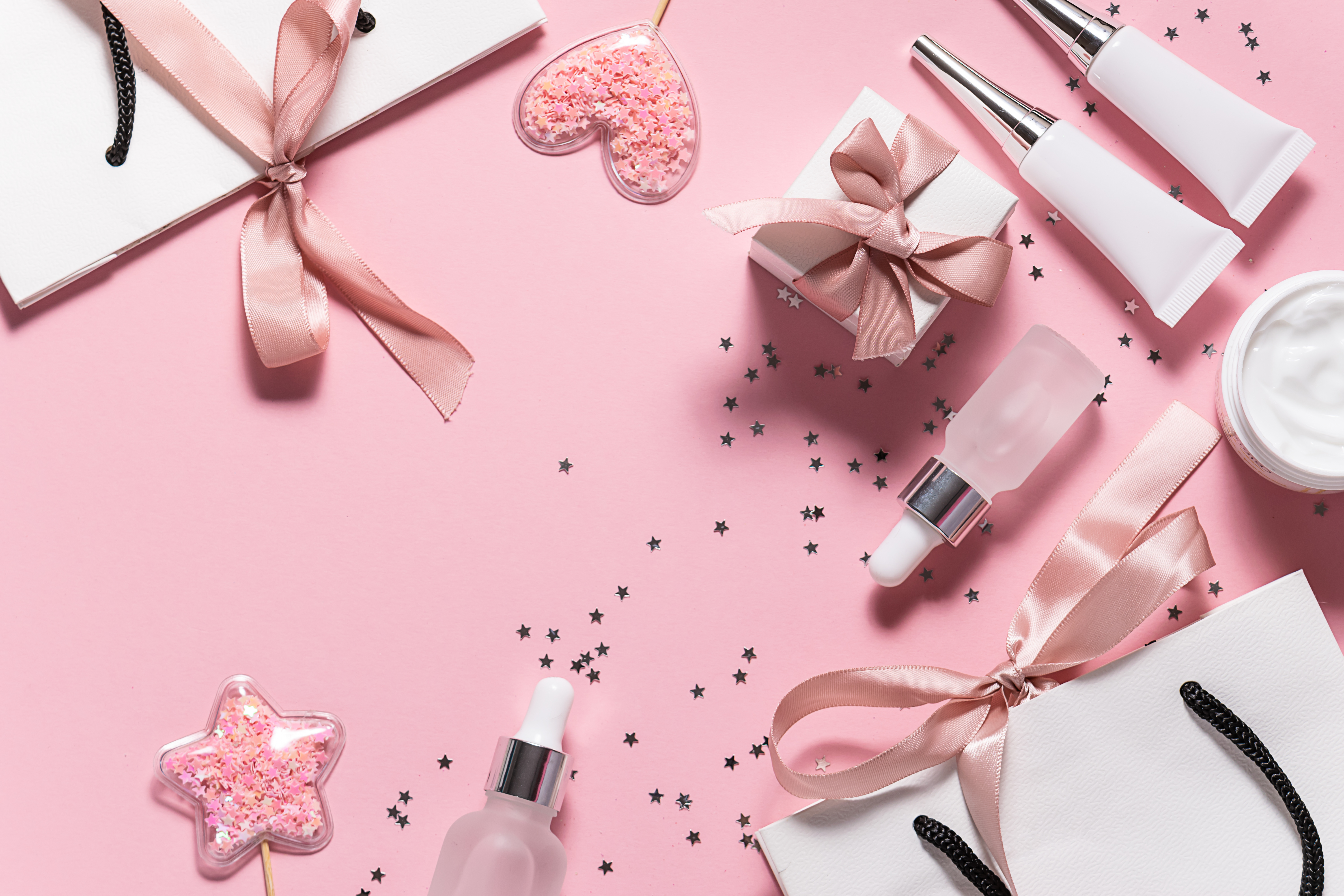 Creative flat lay overhead view of skin care products in unbranded containers, gift boxes and bags with ribbons, shining decorations on pink pastel background. Christmas or Valentines Day background.