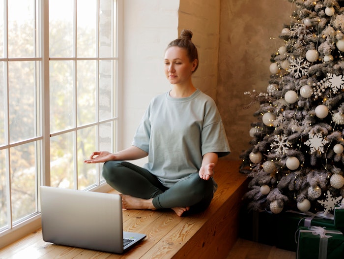 Portrait of young woman meditating while sitting near Christmas tree using laptop for online yoga lessons.