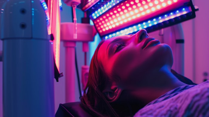 A woman lies on a table with her eyes closed, receiving red light therapy. The red light panel is positioned directly above her head.
