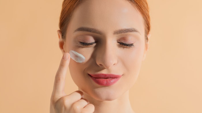 redheaded beautiful woman eyes closed applying soothing lotion to face beige background