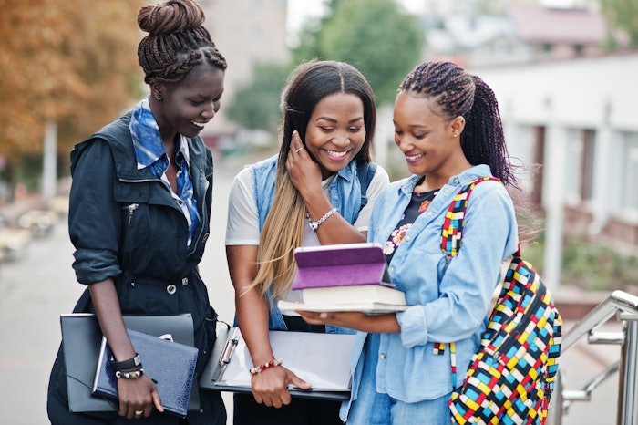 Three Black students on a college campus, carrying books, bags and other school supplies.