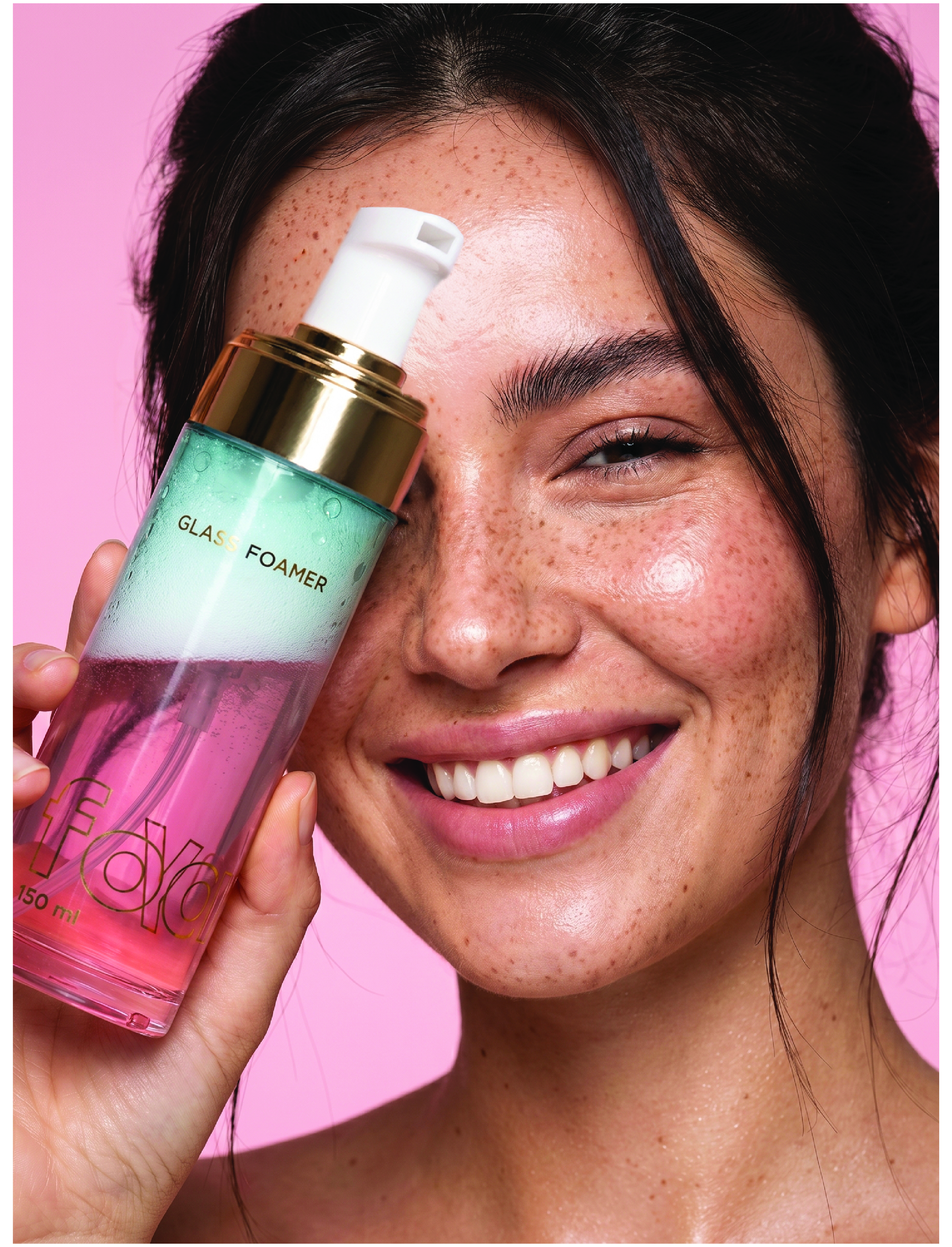Woman holding GLAS FOAMER facial cleanser bottle, smiling against pink background with visible freckles