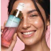 Woman holding GLAS FOAMER facial cleanser bottle, smiling against pink background with visible freckles