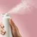 Hand holding white spray bottle dispensing mist against pink background