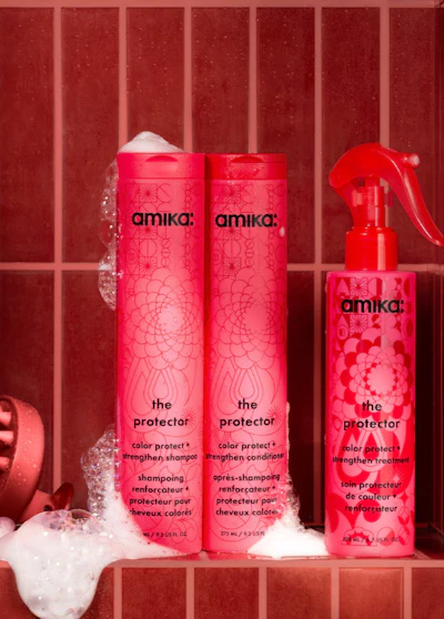 Amika hair care products displayed on red tile background with shampoo, conditioner, and treatment spray bottles