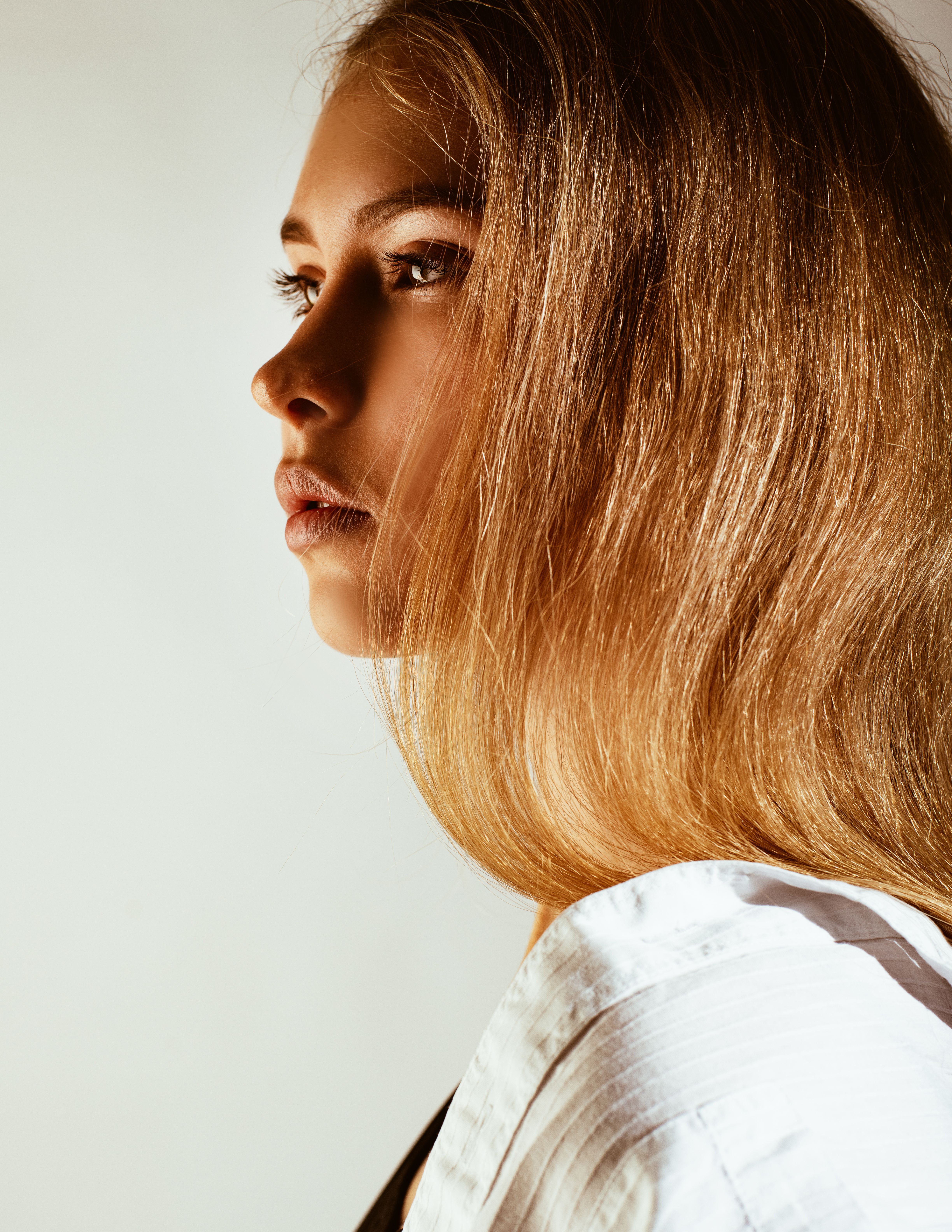 Woman with long caramel-blonde hair in profile wearing white shirt against neutral background
