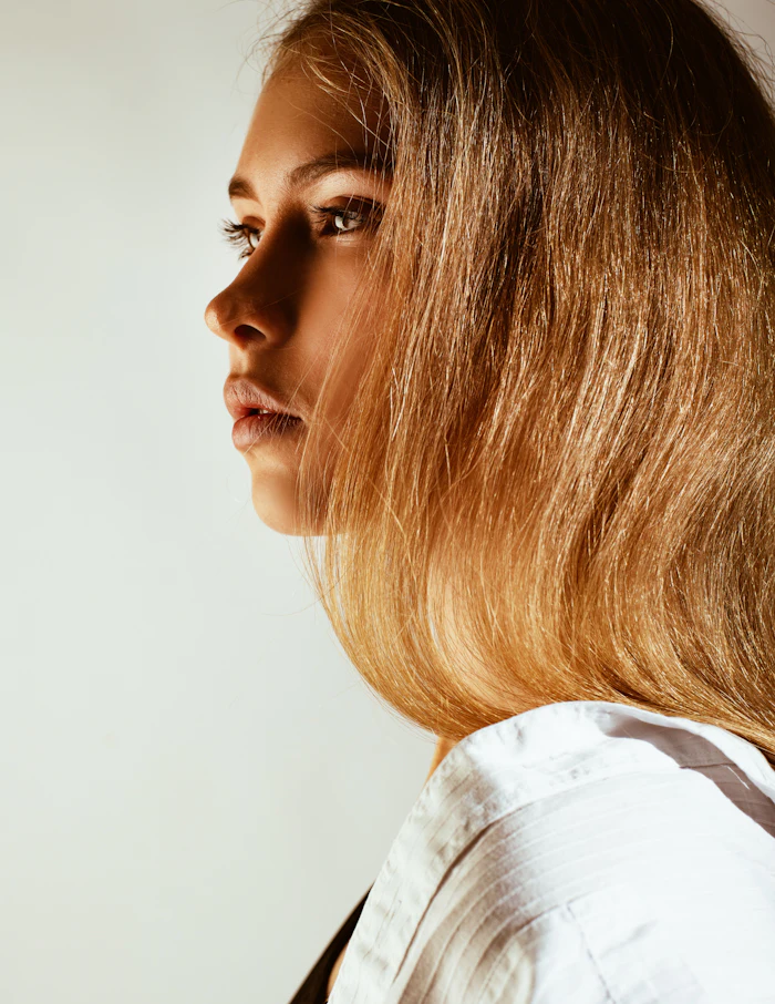 Woman with long caramel-blonde hair in profile wearing white shirt against neutral background