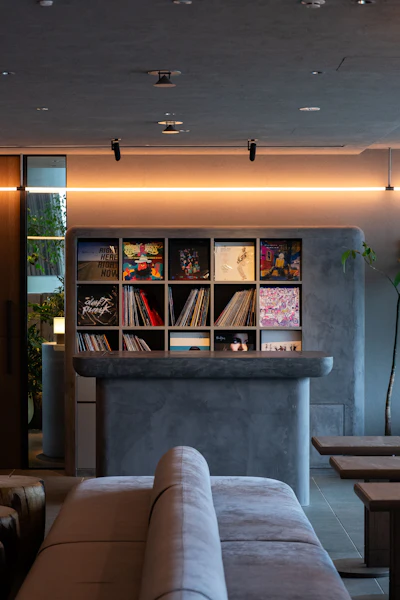 Modern lounge interior with concrete shelving unit displaying vinyl records and artwork, warm ambient lighting, and minimalist furnishings