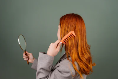 Woman with long orange hair combing her hair while looking in a hand mirror against a gray background