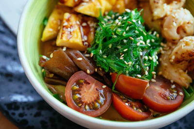 Asian noodle bowl with tofu, shrimp, seaweed, tomato, and sesame seeds in green-rimmed white bowl