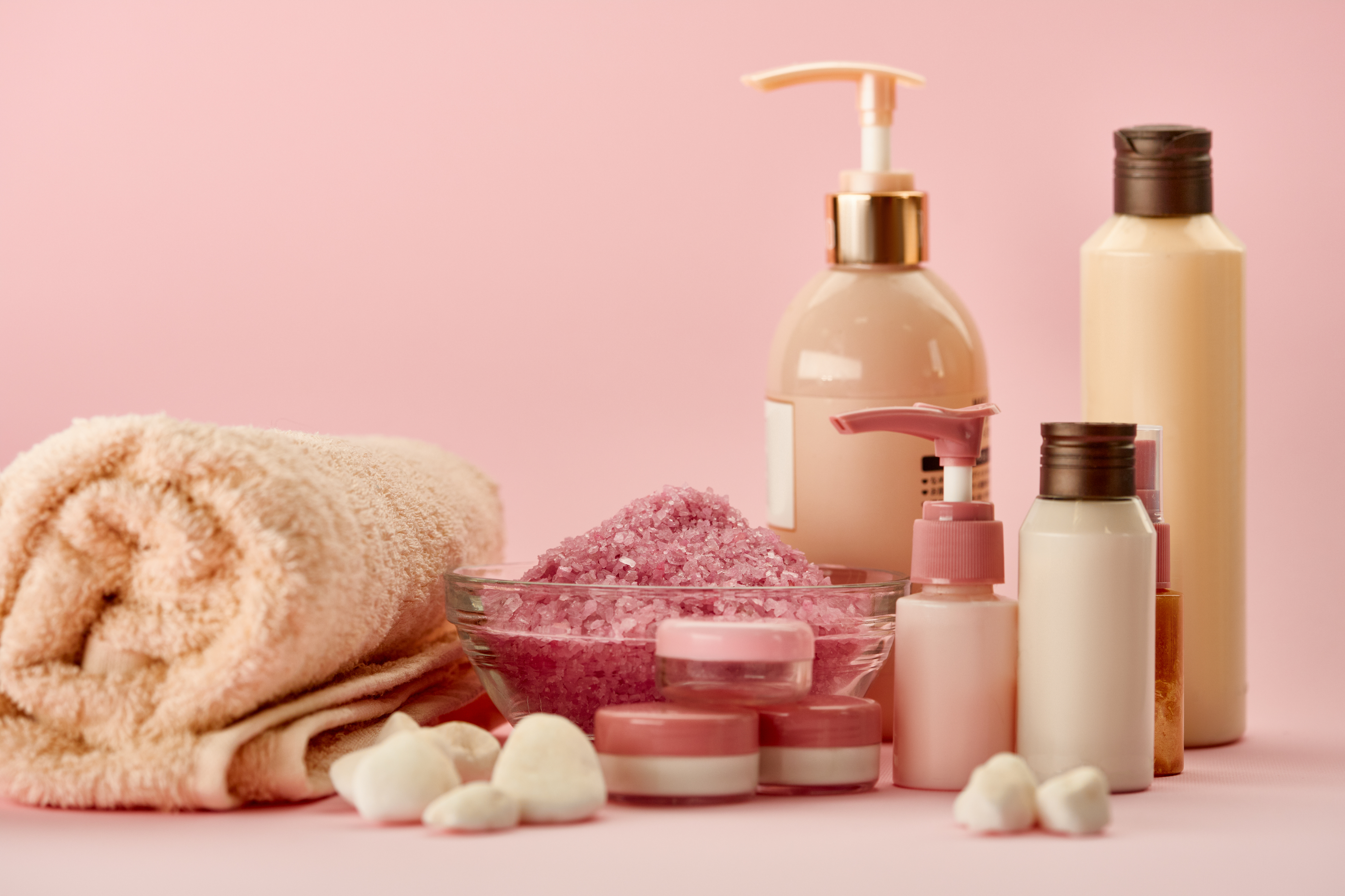 Spa and skincare products including bottles, jars, bath salts, towel, and bath bombs arranged on pink background