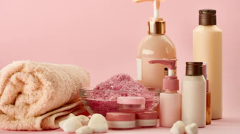 Spa and skincare products including bottles, jars, bath salts, towel, and bath bombs arranged on pink background