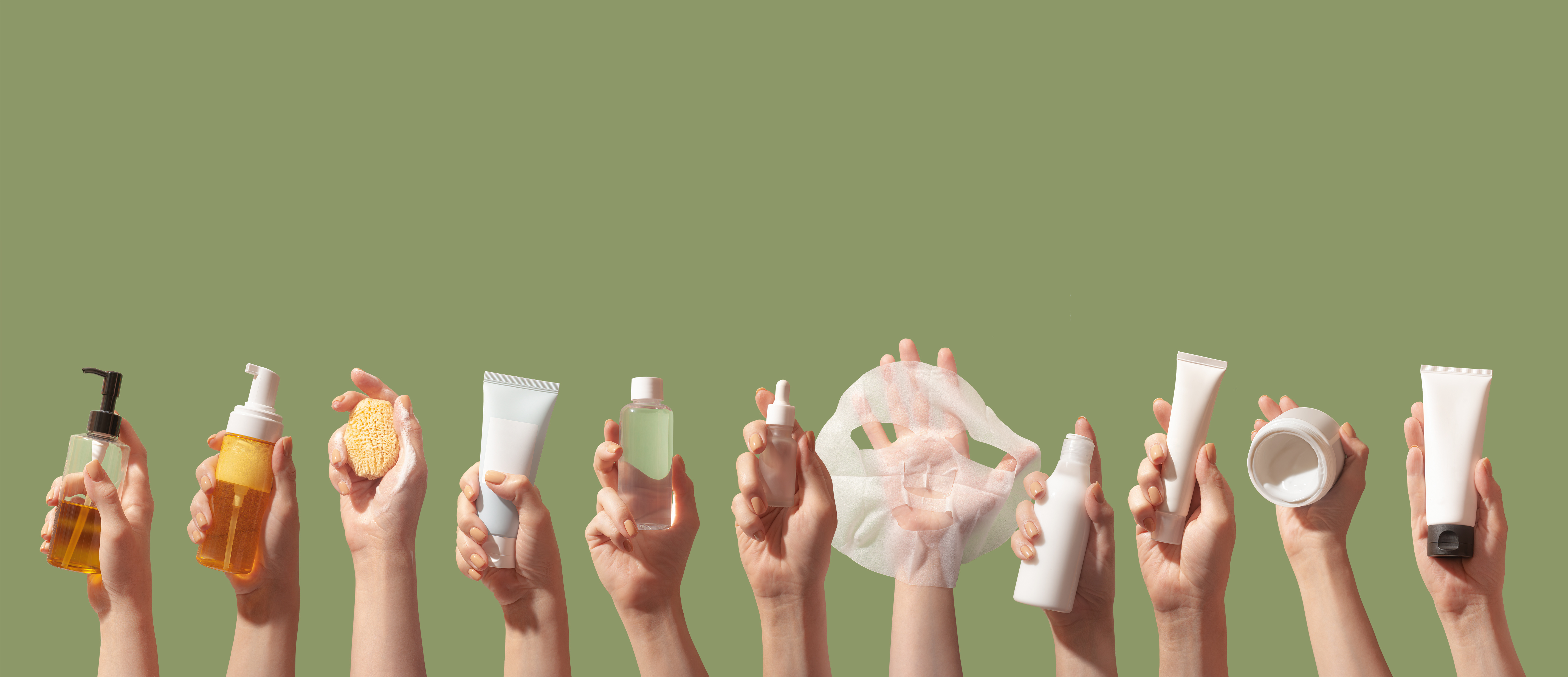 Hands holding assorted beauty and skincare products including serums, creams, and cleansers on green background