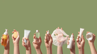 Hands holding assorted beauty and skincare products including serums, creams, and cleansers on green background