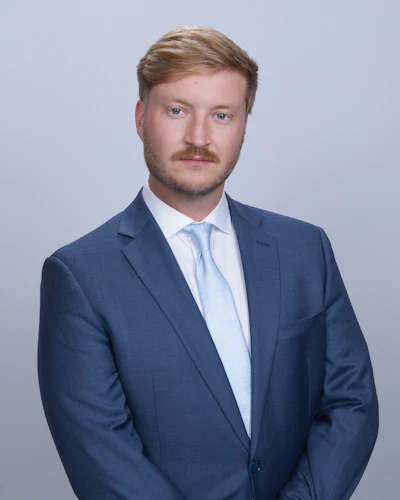 Professional headshot of a man in navy blazer and light blue tie against neutral background