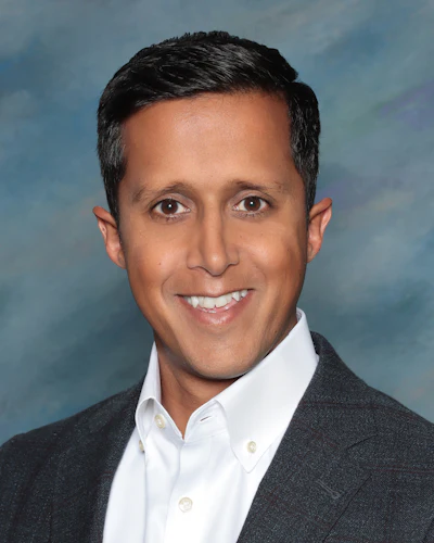 Professional headshot of a man in dark blazer and white shirt smiling against blue background