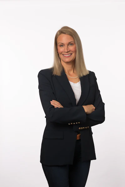 Professional woman in black blazer and pearl necklace smiling with arms crossed against white background