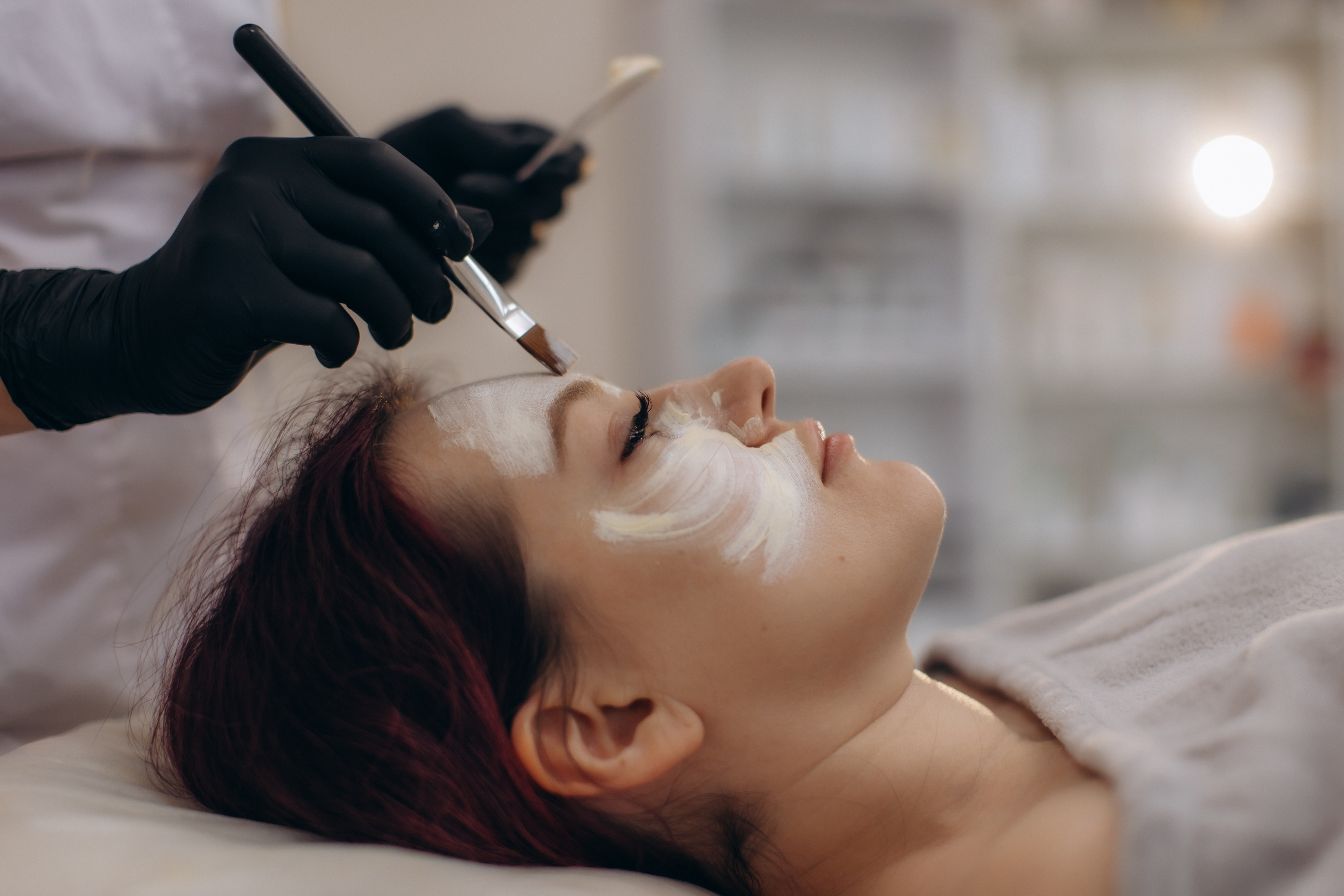 Esthetician applying white facial mask to client during professional skincare treatment
