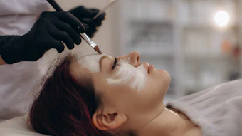 Esthetician applying white facial mask to client during professional skincare treatment