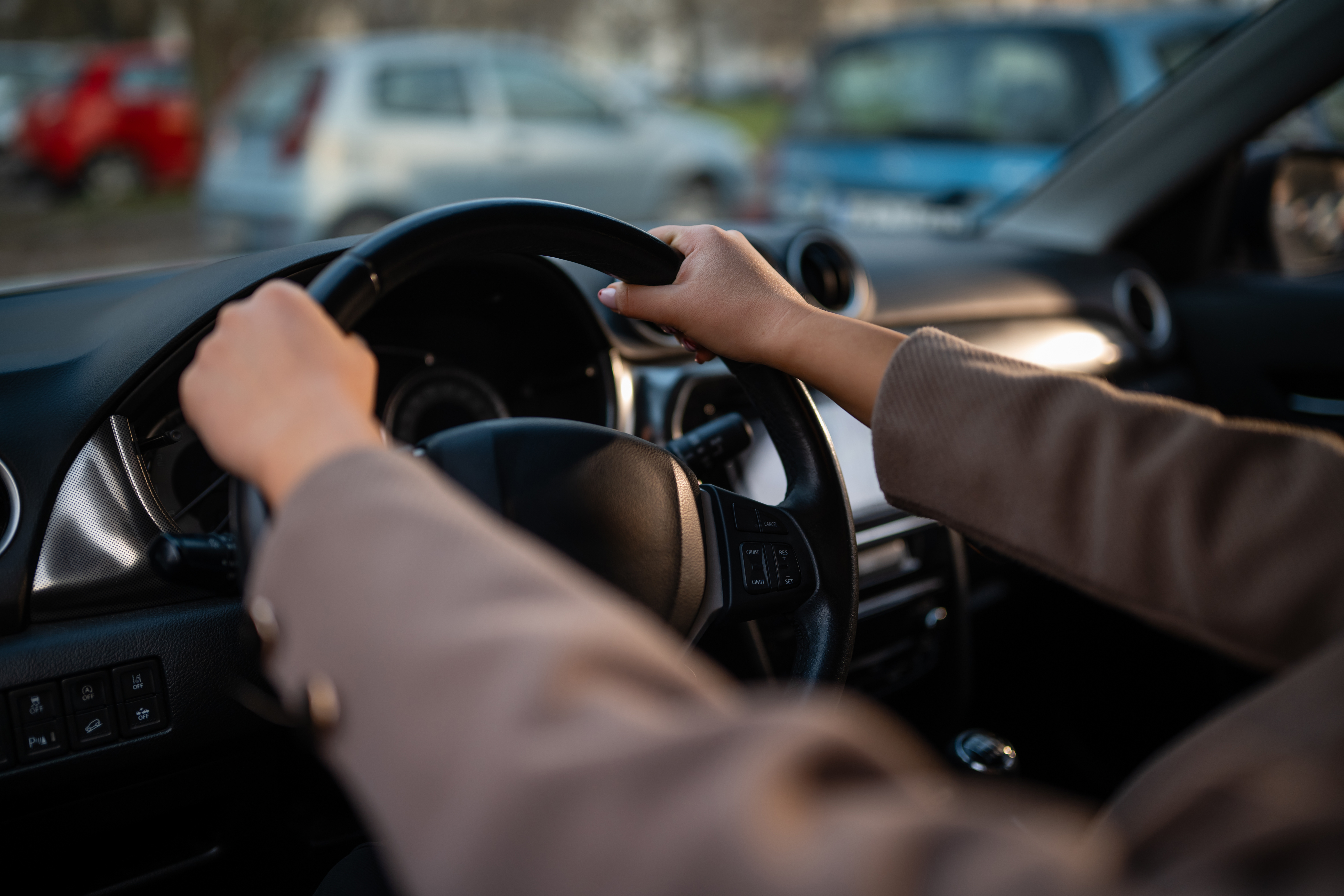 Driver's hands on steering wheel in sunny vehicle interior with parked cars visible through windshield