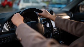 Driver's hands on steering wheel in sunny vehicle interior with parked cars visible through windshield