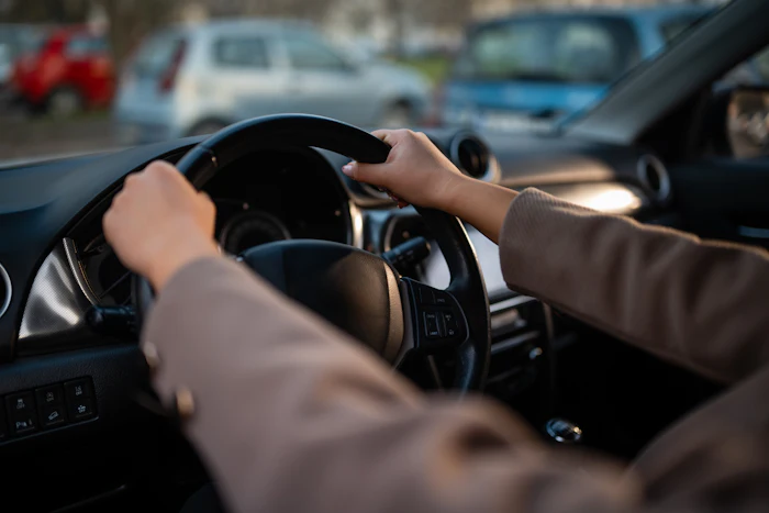 Driver's hands on steering wheel in sunny vehicle interior with parked cars visible through windshield