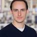 Professional headshot of a man with dark hair wearing a navy sweater in an office setting