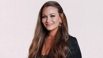 Professional headshot of a smiling woman with long brown hair wearing a black blazer and gold jewelry