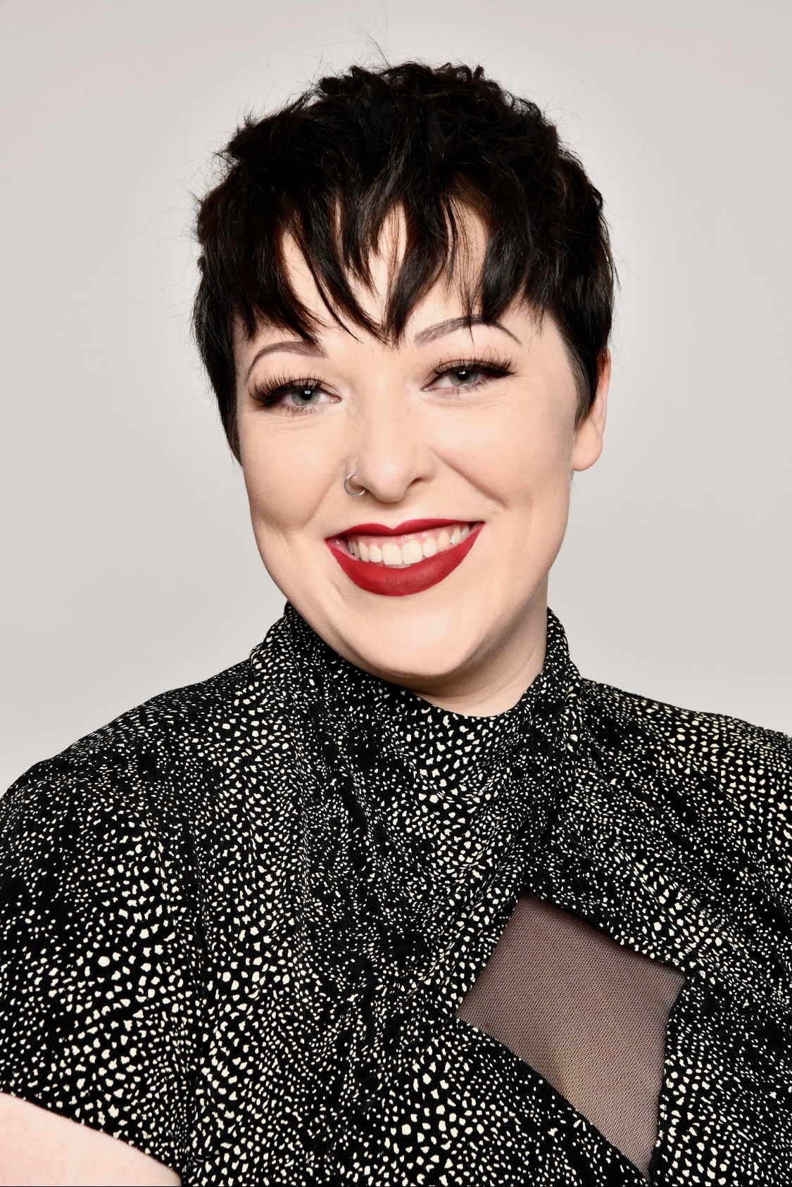 Professional headshot of a woman with short black hair, red lipstick, wearing a black and white patterned jacket against a neutral background