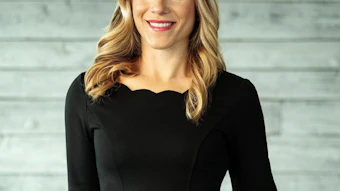 Professional headshot of a woman with blonde wavy hair wearing a black short-sleeved dress against a light brick background