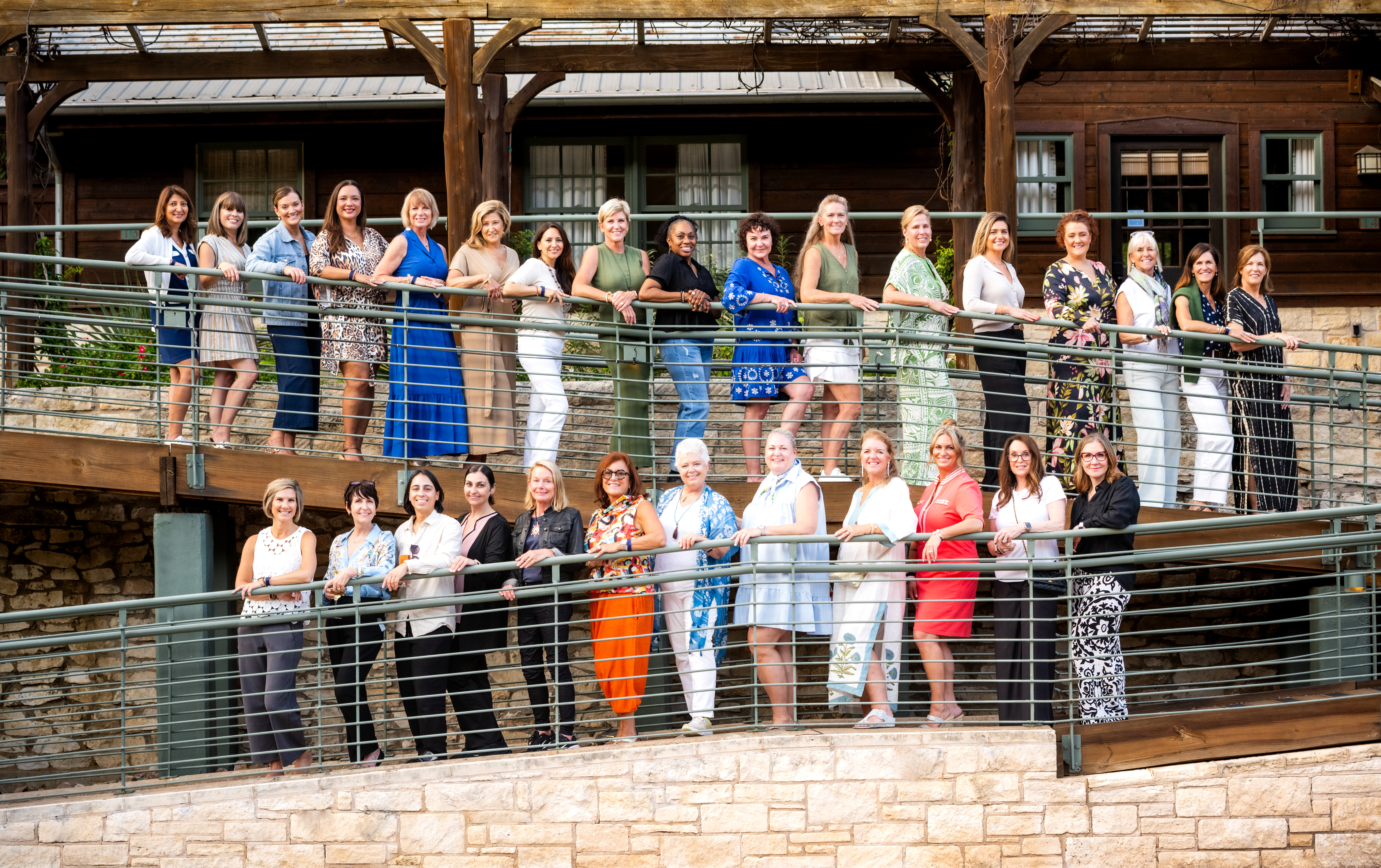 Large group of women posed together on a two-level wooden deck with metal railings at a rustic venue