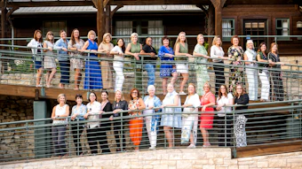 Large group of women posed together on a two-level wooden deck with metal railings at a rustic venue